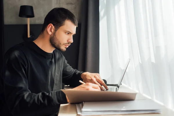 Foco seletivo de freelancer barbudo usando laptop e segurando lápis perto da pasta em casa — Fotografia de Stock