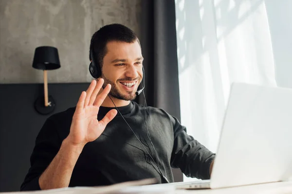 Enfoque selectivo de operador feliz en auriculares saludando mano mientras tiene videollamada en casa - foto de stock