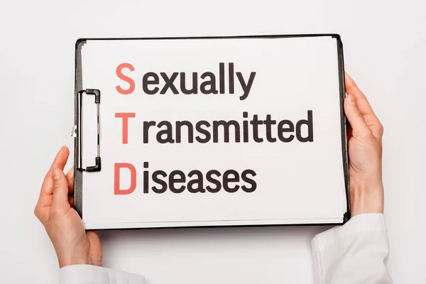 Vista recortada del médico sujetando portapapeles con letras de enfermedades de transmisión sexual aisladas en blanco - foto de stock