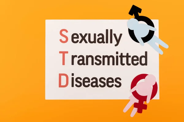 Visão superior de pessoas de papel com símbolos de gênero perto de papel com doenças sexualmente transmissíveis lettering isolado em laranja — Fotografia de Stock