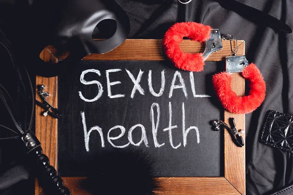 Top view of mask near chalkboard with sexual health lettering and sex toys on black silk — Stock Photo