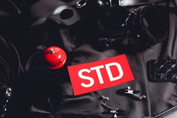 Top view of red sex bell near paper with std lettering and sex toys on black — Stock Photo