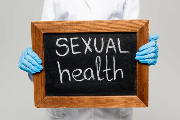 Vue recadrée du médecin dans des gants en latex tenant tableau avec lettrage de santé sexuelle isolé sur gris — Photo de stock