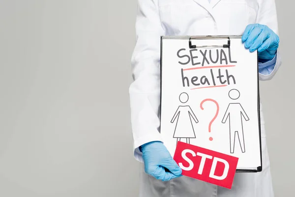 Vista recortada del médico en guantes de látex azul y capa blanca sujetando portapapeles con salud sexual, dibujado hombre y mujer y std letras aisladas en gris - foto de stock