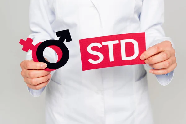 Cropped view of doctor in white coat holding gender symbols and std lettering isolated on grey — Stock Photo
