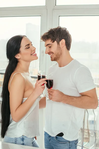 Hermosa pareja sonriente sosteniendo vasos con vino tinto durante el auto aislamiento cerca de la ventana - foto de stock