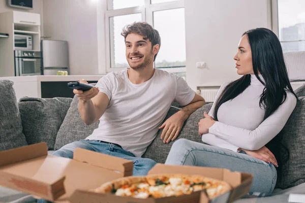 Эмоциональная пара, которая ест пиццу и смотрит телевизор с пультом дистанционного управления во время самоизоляции дома — стоковое фото