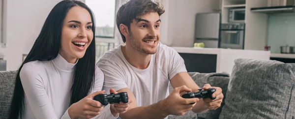 KYIV, UCRANIA - 16 DE ABRIL DE 2020: hermosa pareja alegre jugando videojuegos con joysticks durante el autoaislamiento en casa, encabezado del sitio web - foto de stock