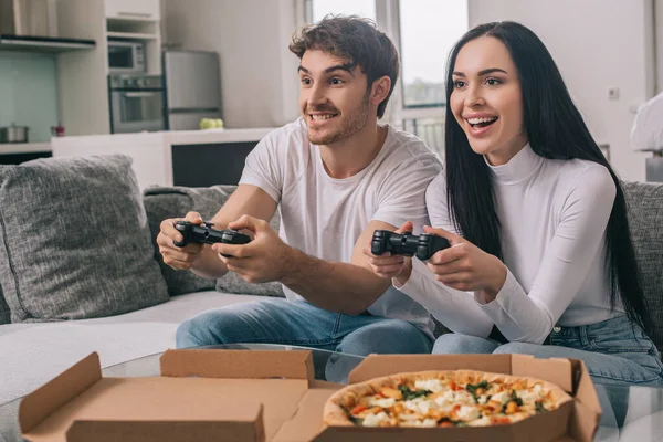 KYIV, UKRAINE - 16 de abril de 2020: casal alegre tendo pizza e jogando videogame com joysticks durante o auto-isolamento em casa — Fotografia de Stock