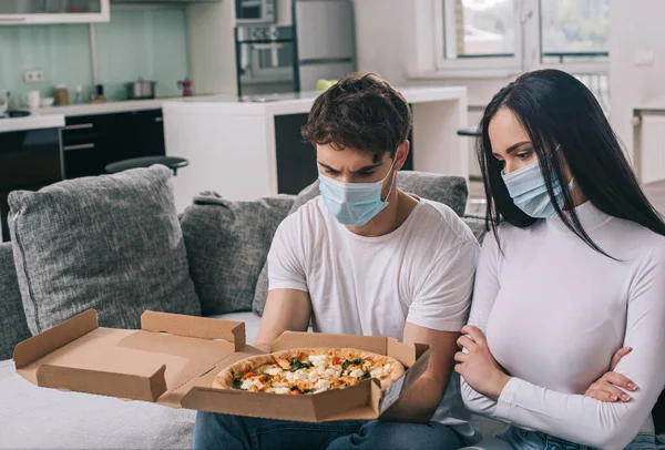 Pareja enferma en máscaras médicas mirando pizza en casa durante el auto aislamiento - foto de stock