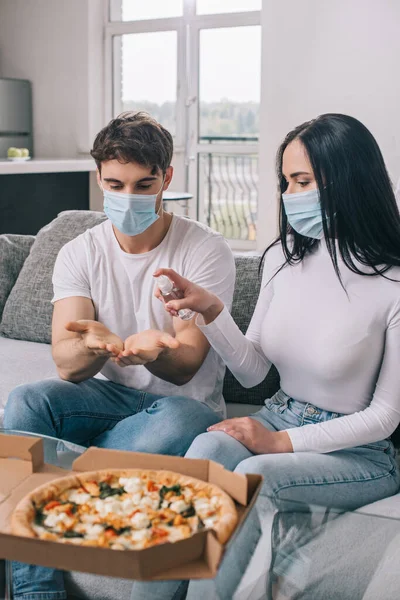 Pareja enferma en máscaras médicas usando aerosol antiséptico antes de comer pizza en casa durante el auto aislamiento - foto de stock