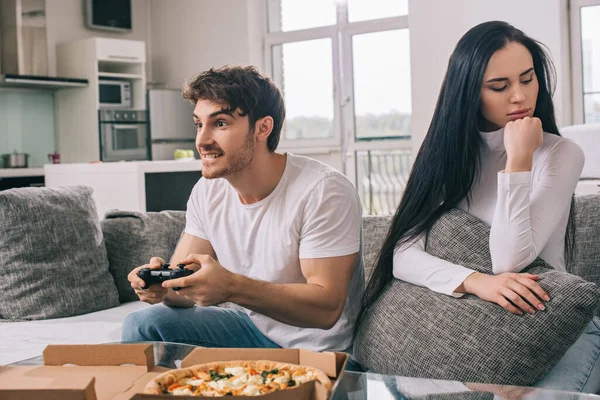 KYIV, UKRAINE - APRIL 16, 2020: offended woman sitting near concentrated man playing video game with joystick during self isolation at home with pizza — Stock Photo