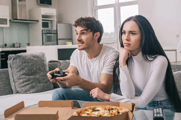 KYIV, UCRANIA - 16 de ABRIL de 2020: chica triste sentada cerca de novio jugando videojuego con joystick durante el autoaislamiento en casa con pizza - foto de stock