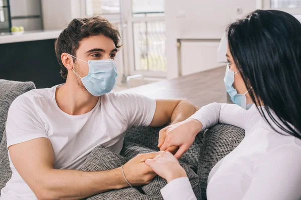 Ill couple in medical masks holding hands at home during self isolation — Stock Photo