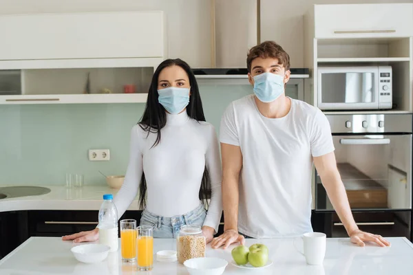 Pareja en máscaras médicas desayunando en casa durante el autoaislamiento - foto de stock