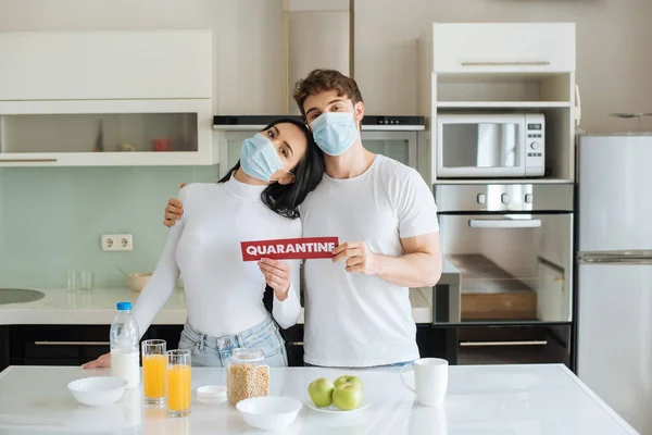 Paar in medizinischen Masken frühstückt zu Hause und hält Quarantäne-Zeichen während der Selbstisolierung — Stock Photo