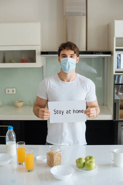 Ill man in medical mask holding stay at home card on kitchen during breakfast on self isolation — Stock Photo