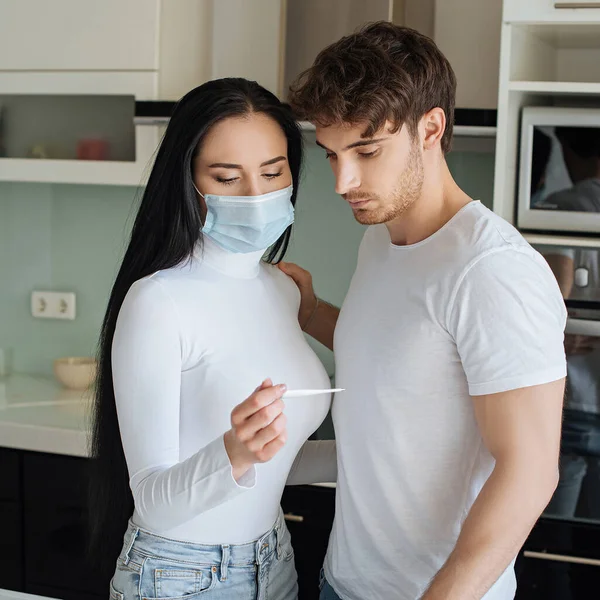 Novio y chica enferma en máscara médica mirando el termómetro durante el auto aislamiento - foto de stock