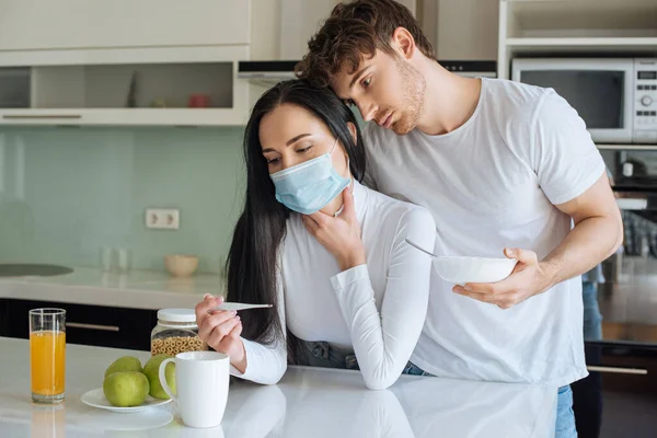 Homme et femme malade en masque médical regardant le thermomètre et prenant le petit déjeuner pendant l'isolement personnel — Photo de stock