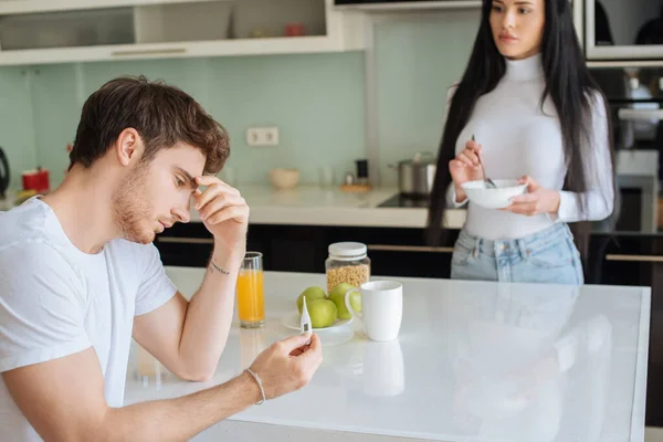 Ill man holding thermometer while woman holding bowl with breakfast at home during self isolation, selective focus — Stock Photo