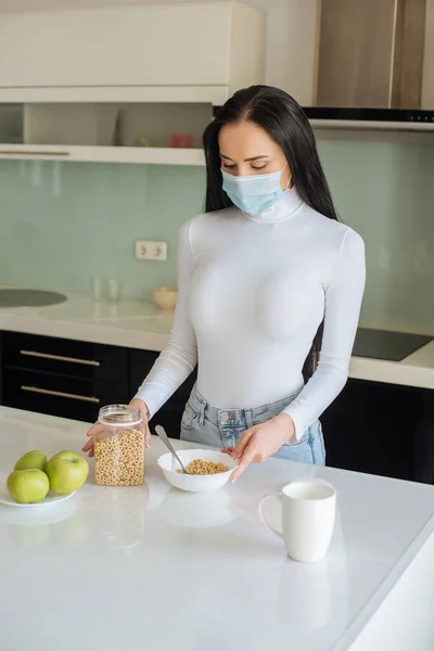 Traurige Frau in medizinischer Maske mit Blick auf Cornflakes und Äpfel zum Frühstück während der Selbstisolierung zu Hause — Stockfoto