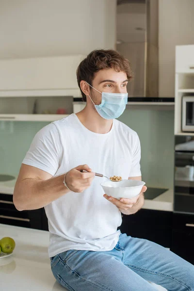 Homem doente bonito em tigela máscara médica segurando com flocos de milho na cozinha em auto-isolamento — Fotografia de Stock