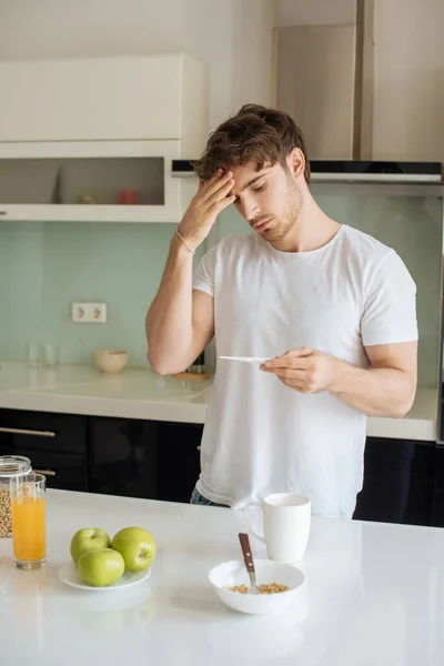 Preocupado hombre enfermo con fiebre mirando termómetro en la cocina durante el desayuno en auto aislamiento - foto de stock