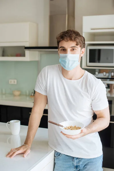 Enfermo hombre en médico máscara celebración bowl con cornflakes en cocina en auto aislamiento - foto de stock