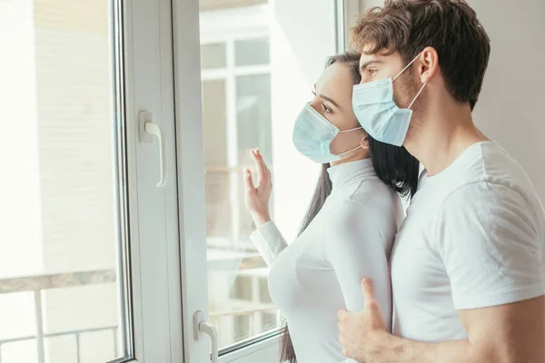 Ill couple in medical masks looking through window during self isolation — Stock Photo