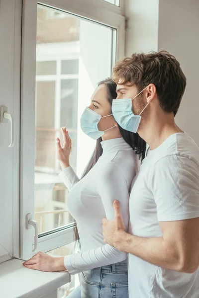 Красивая пара в медицинских масках, смотрящая в окно во время самоизоляции — стоковое фото