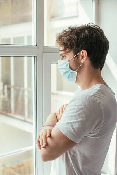 Молодий сумний чоловік у медичній масці з схрещеними руками дивиться крізь вікно під час самоізоляції — стокове фото