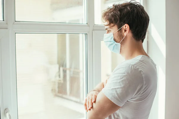 Triste homem doente em máscara médica com braços cruzados olhando através da janela durante o auto-isolamento — Fotografia de Stock