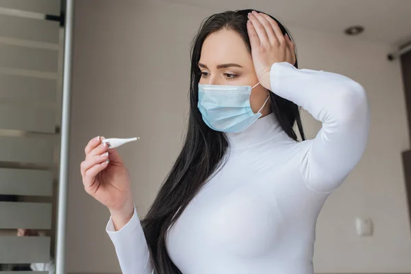 Mujer preocupada con máscara médica con fiebre mirando el termómetro durante el autoaislamiento en casa - foto de stock