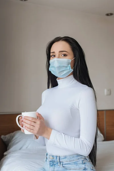 Worried woman in medical mask holding cup of tea in bedroom during self isolation — Stock Photo