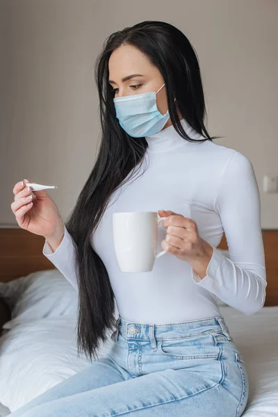 Donna preoccupata in maschera medica in possesso di tazza e termometro durante l'auto isolamento a casa — Foto stock