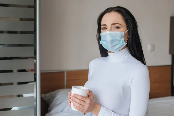 Ill woman in medical mask holding cup of hot drink during self isolation at home — Stock Photo