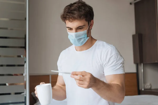 Kranker Mann in medizinischer Maske hält Thermometer und Tasse mit Heißgetränk während der Selbstisolation — Stockfoto