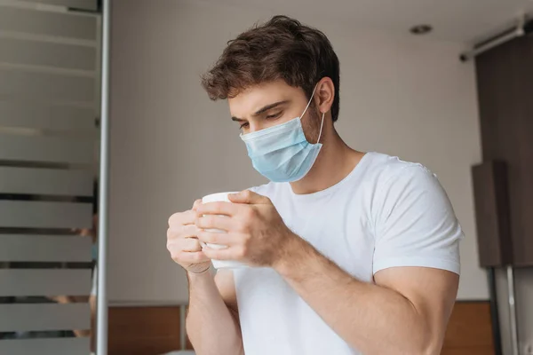 Hombre enfermo en máscara médica mirando taza con bebida caliente en el dormitorio durante el aislamiento del uno mismo - foto de stock