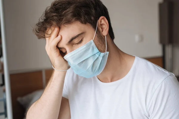 Erschöpfter kranker Mann in medizinischer Maske sitzt während Selbstisolierung im Schlafzimmer — Stockfoto