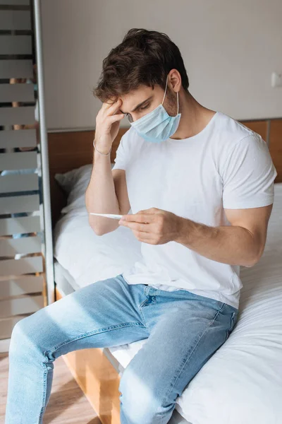 Hombre enfermo cansado en máscara médica mirando el termómetro durante el auto aislamiento - foto de stock