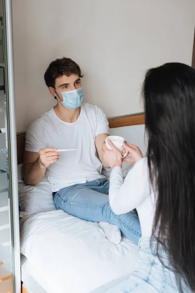 Woman giving cup of hot drink to ill man in medical mask with thermometer in bed during self isolation — Stock Photo