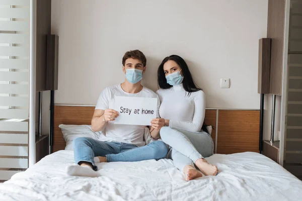 Joven pareja en médico máscaras holding estancia en casa signo en dormitorio durante auto aislamiento - foto de stock