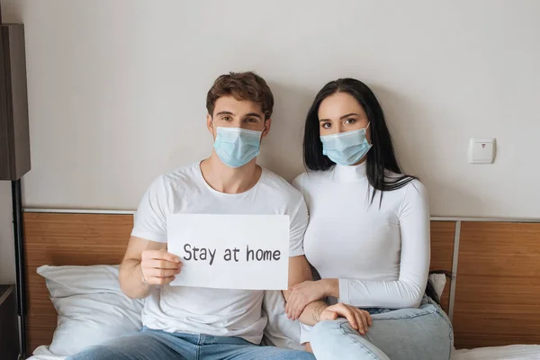 Couple malade dans les masques médicaux tenant rester à la maison signe dans le lit pendant l'isolement personnel — Photo de stock