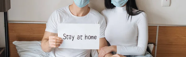 Cropped view of ill couple in medical masks holding Stay at home sign at home during self isolation, horizontal crop — Stock Photo