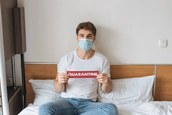 Молодой человек в медицинской маске держит карточку с карантинным словом, сидя на кровати во время самоизоляции — стоковое фото