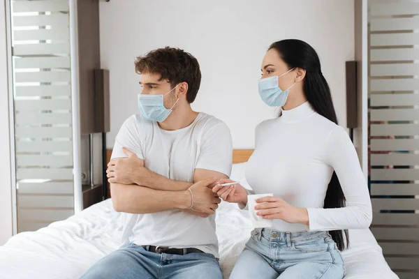 Подружка держит чашку и термометр рядом с больным парнем в медицинской маске на кровати во время самоизоляции — стоковое фото