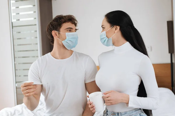 Couple malade dans des masques médicaux tenant tasse et thermomètre et se regardant pendant l'isolement personnel — Photo de stock
