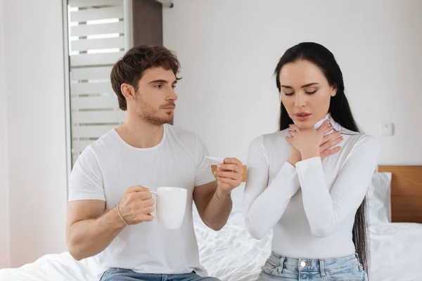 Чоловік дає термометр і чашку гарячого напою хворій жінці з болем в горлі під час самоізоляції — стокове фото