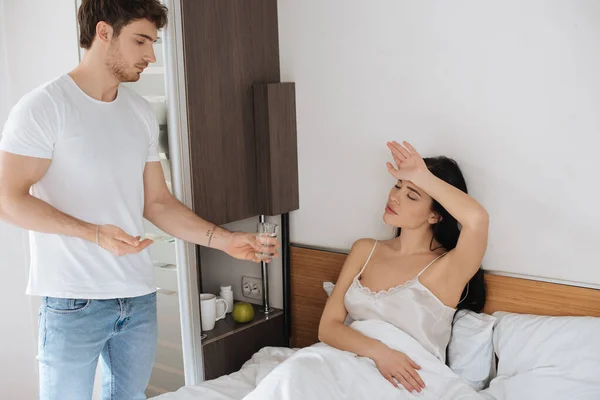 Freund gibt müder kranker Freundin mit Kopfschmerzen während der Selbstisolierung Wasser und Tablette im Schlafzimmer — Stockfoto