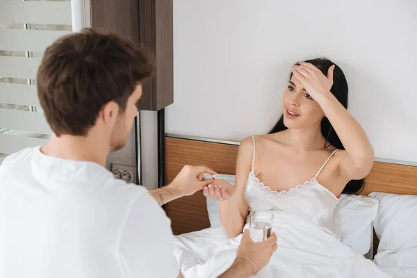 Mann gibt krankes Mädchen mit Kopfschmerzen während der Selbstisolierung Wasser und Tabletten im Bett — Stockfoto